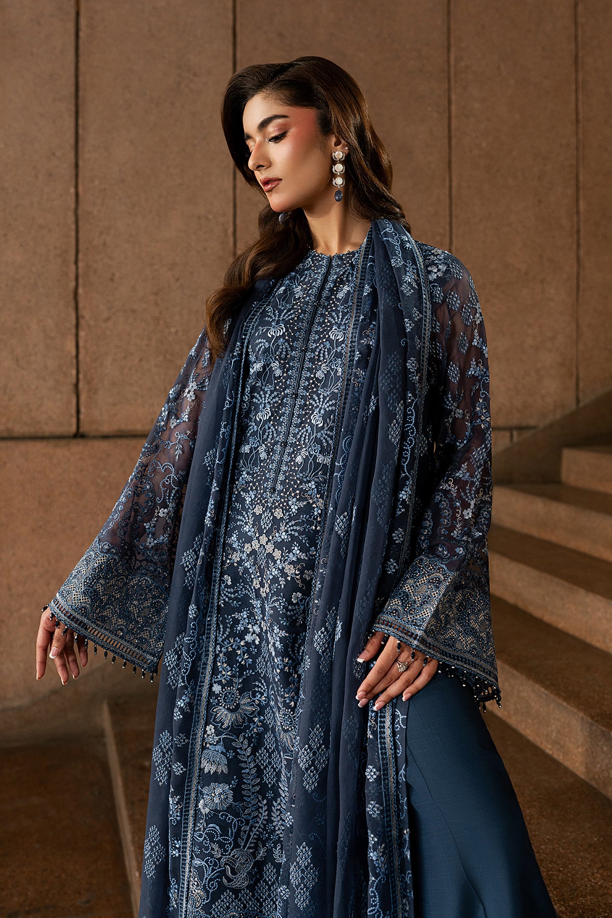 Woman in a blue embroidered outfit standing on a staircase with a beige wall background