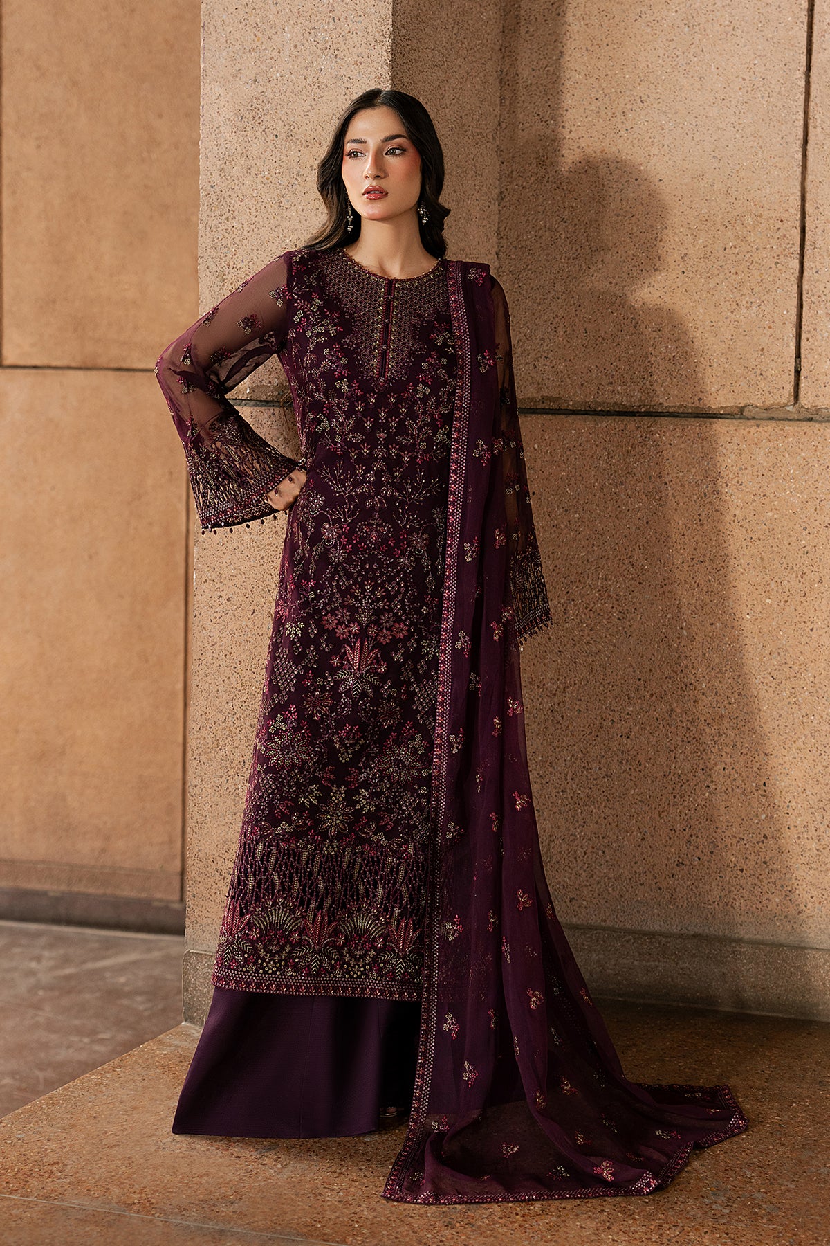 Woman wearing a dark purple embroidered traditional outfit against a beige wall.
