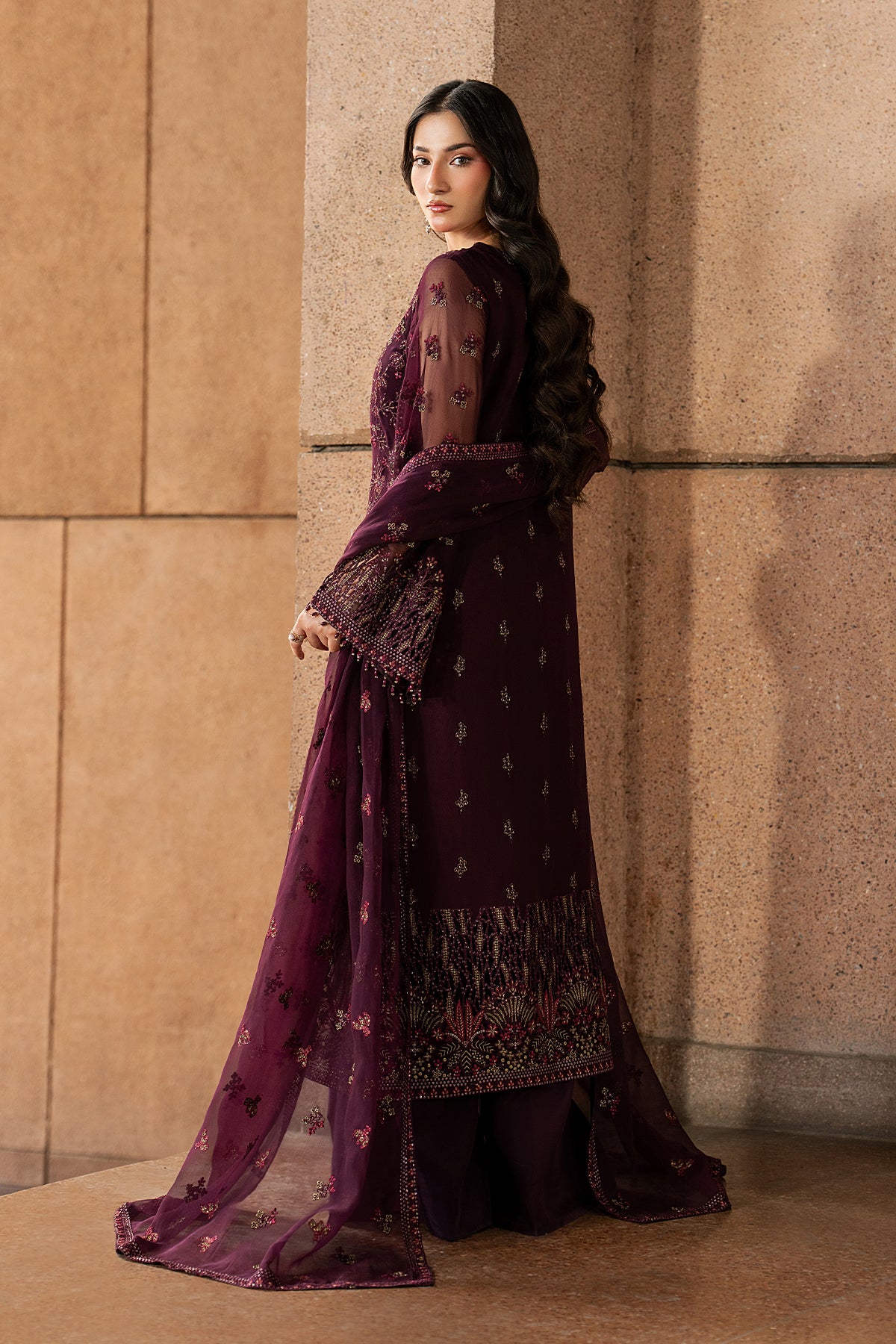 Woman in a dark purple traditional outfit standing against a beige wall.