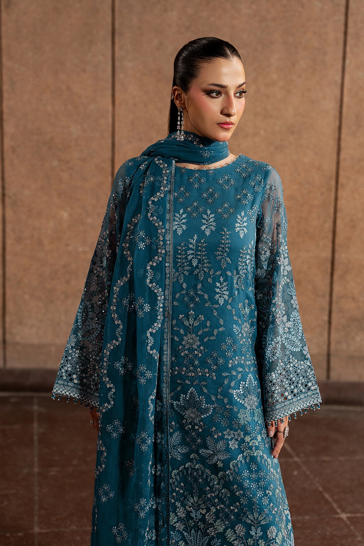Woman wearing a teal embroidered traditional outfit against a brown background