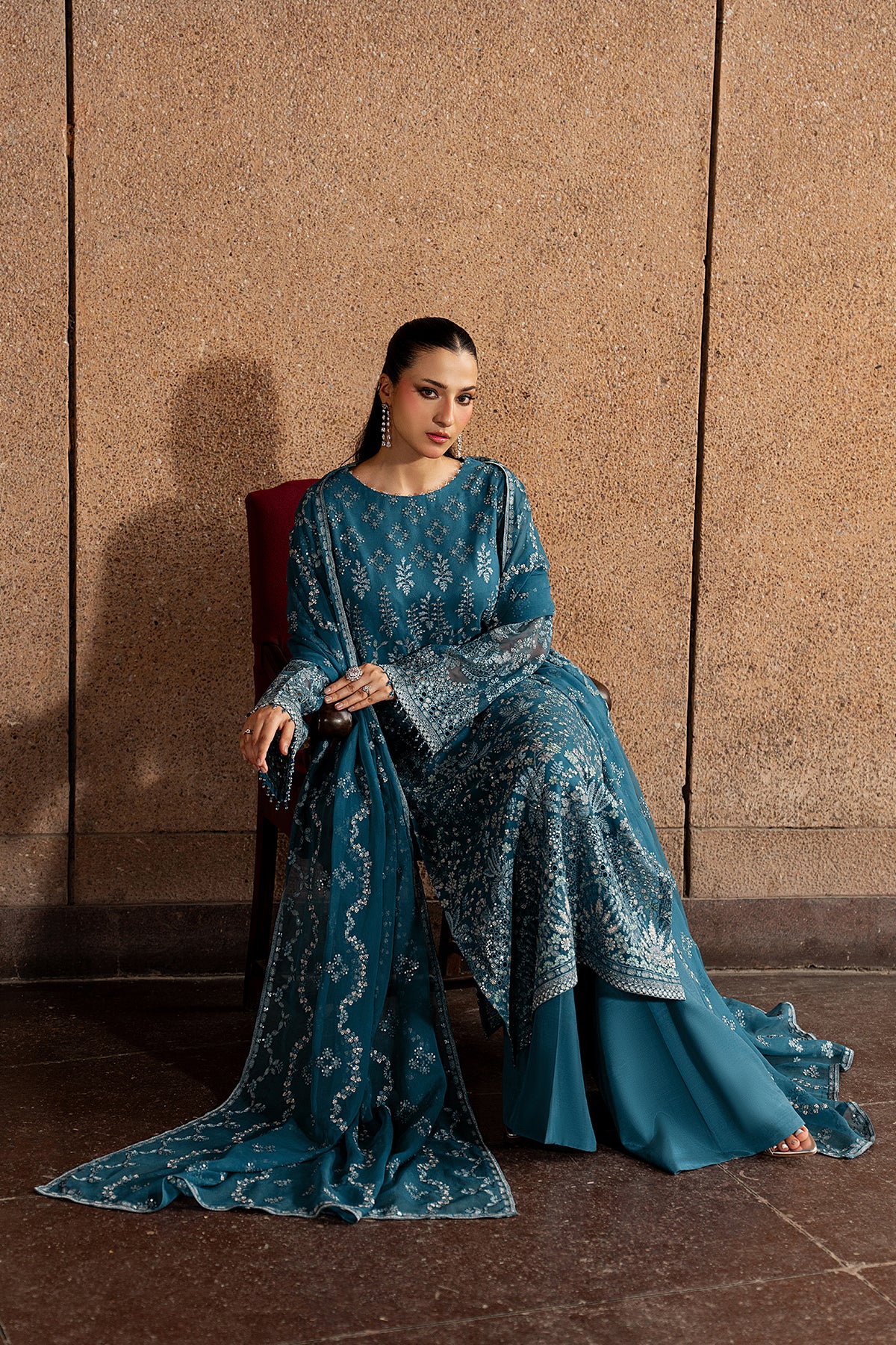 Woman in a blue embroidered outfit sitting against a textured wall.