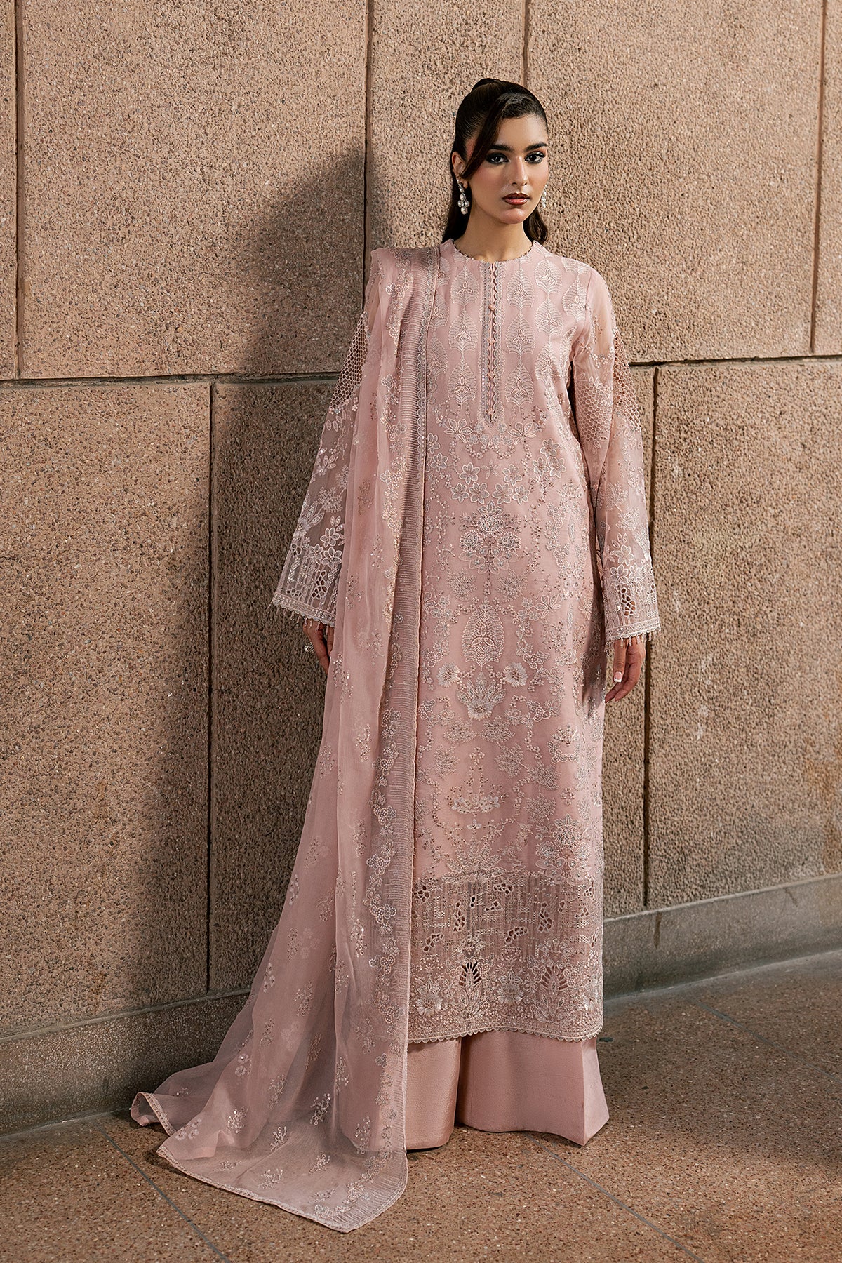 Woman wearing a light pink embroidered outfit against a textured wall.