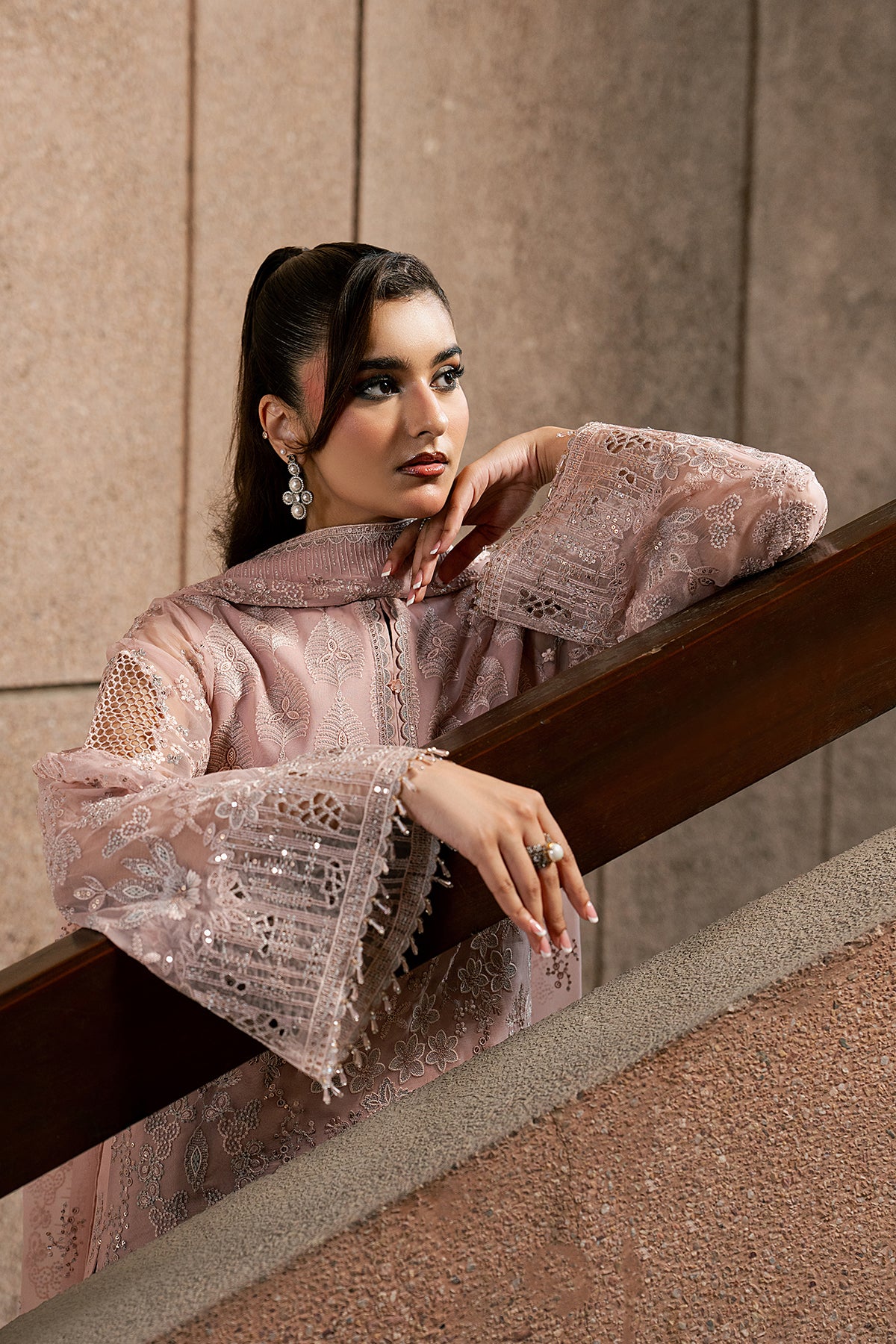 Woman in a pink embroidered outfit leaning against a railing with a neutral wall background