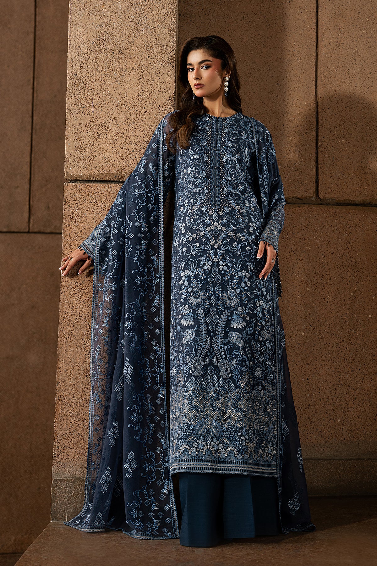 Woman wearing a blue embroidered traditional outfit against a brown stone wall.