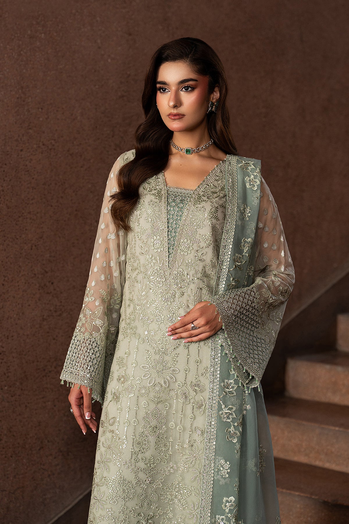 Woman in a light green embroidered traditional outfit standing against a brown background.
