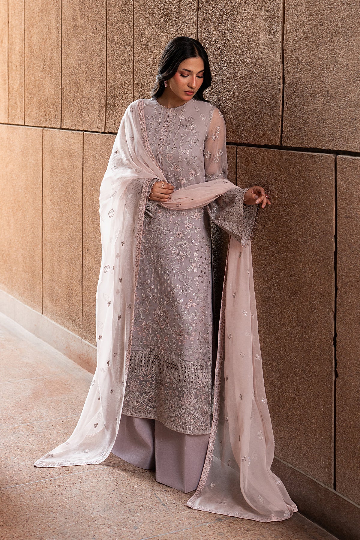 Woman in a light gray traditional outfit with a sheer dupatta against a textured wall.