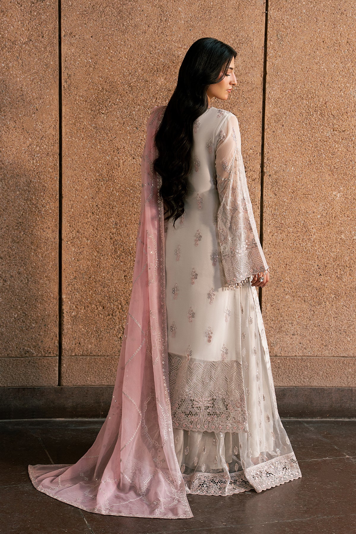 Woman in a traditional outfit with a pink dupatta against a brown wall.