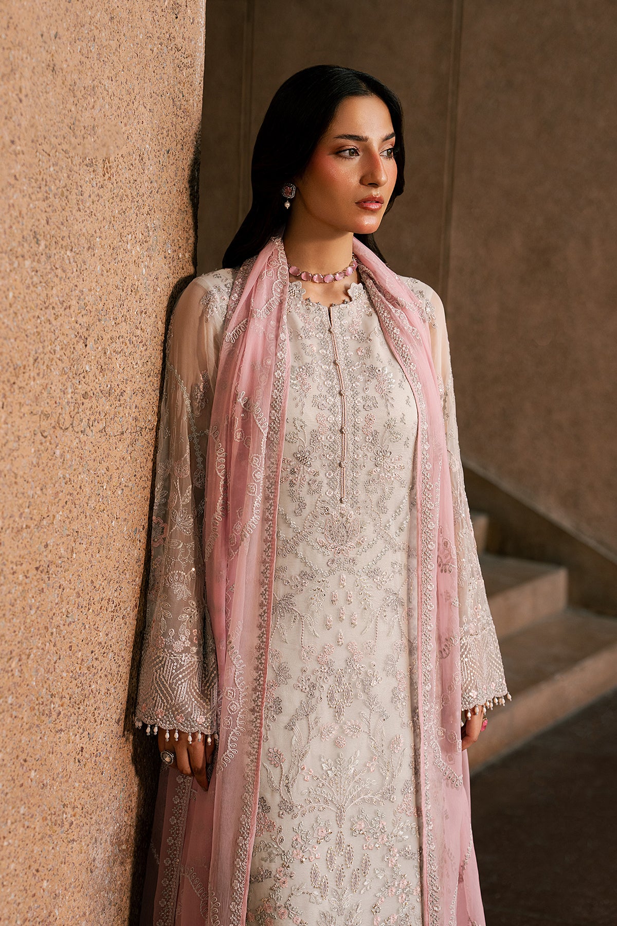 Woman in traditional outfit with pink dupatta leaning against a textured wall.