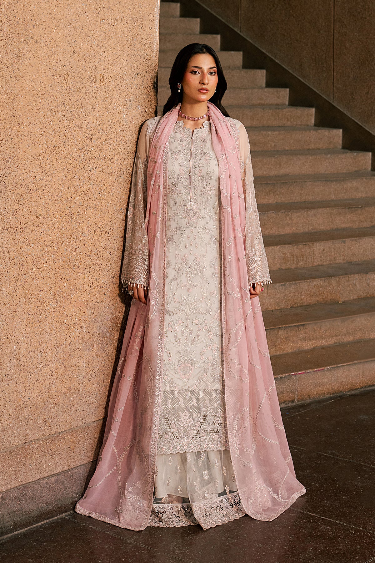 Woman in traditional outfit with pink dupatta standing on a staircase.