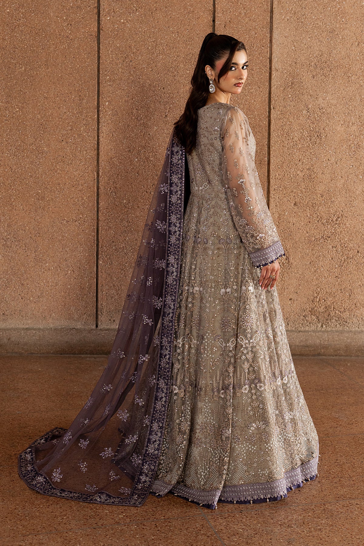 Woman in a traditional outfit with a long, dark dupatta against a beige wall.