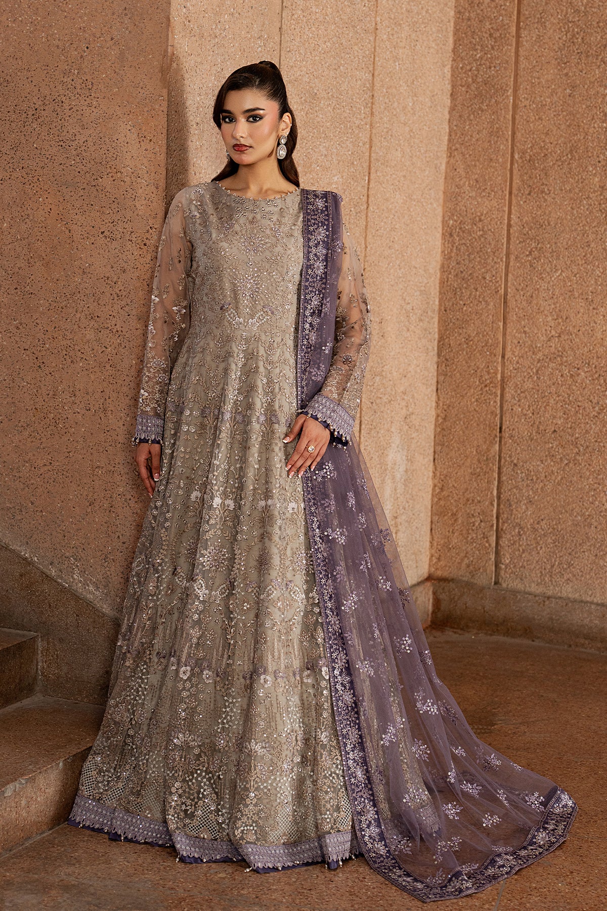 Woman wearing a silver and purple embroidered traditional outfit against a beige wall.