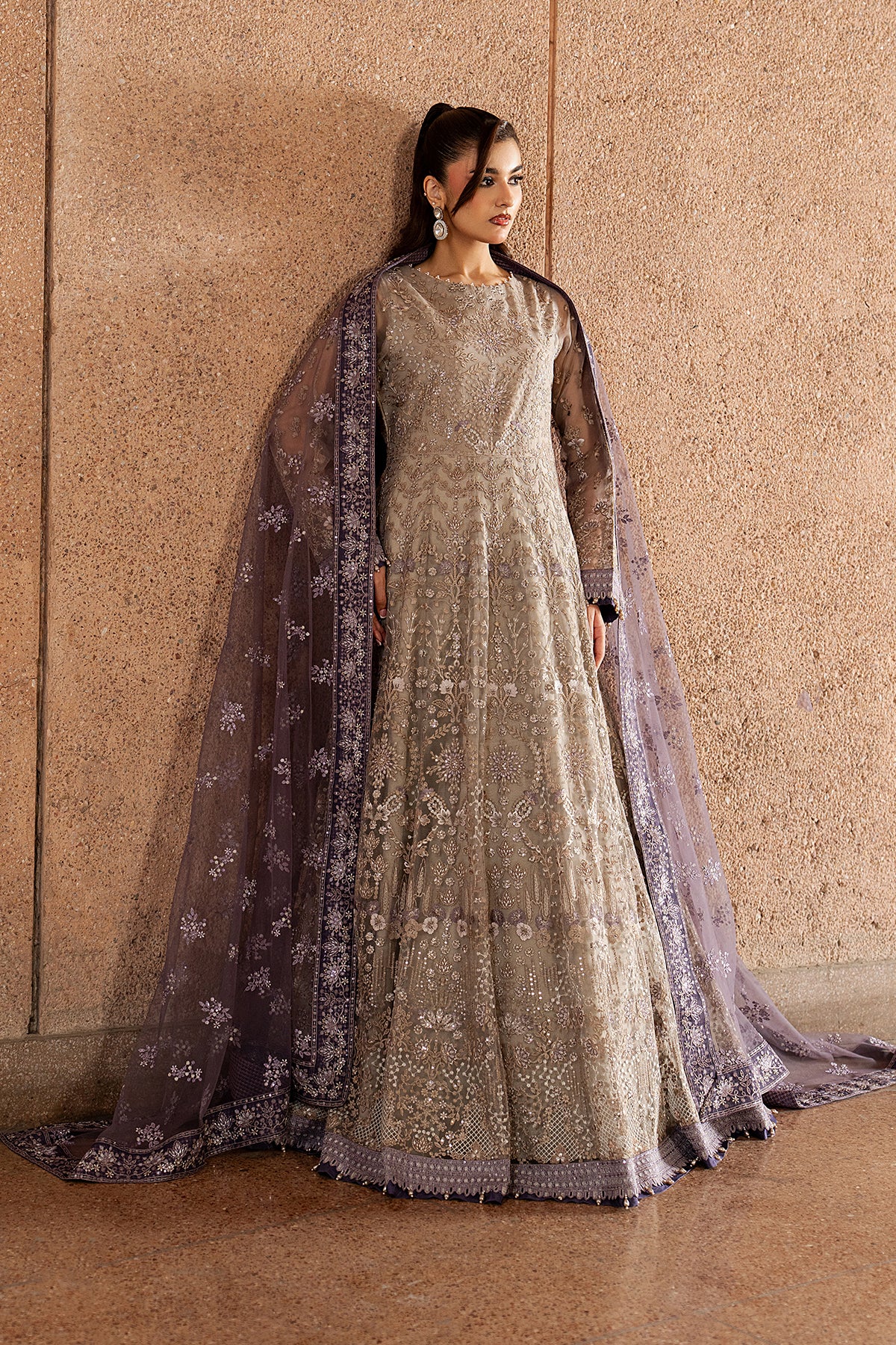 Woman in an elegant embroidered dress with a matching dupatta against a beige wall.
