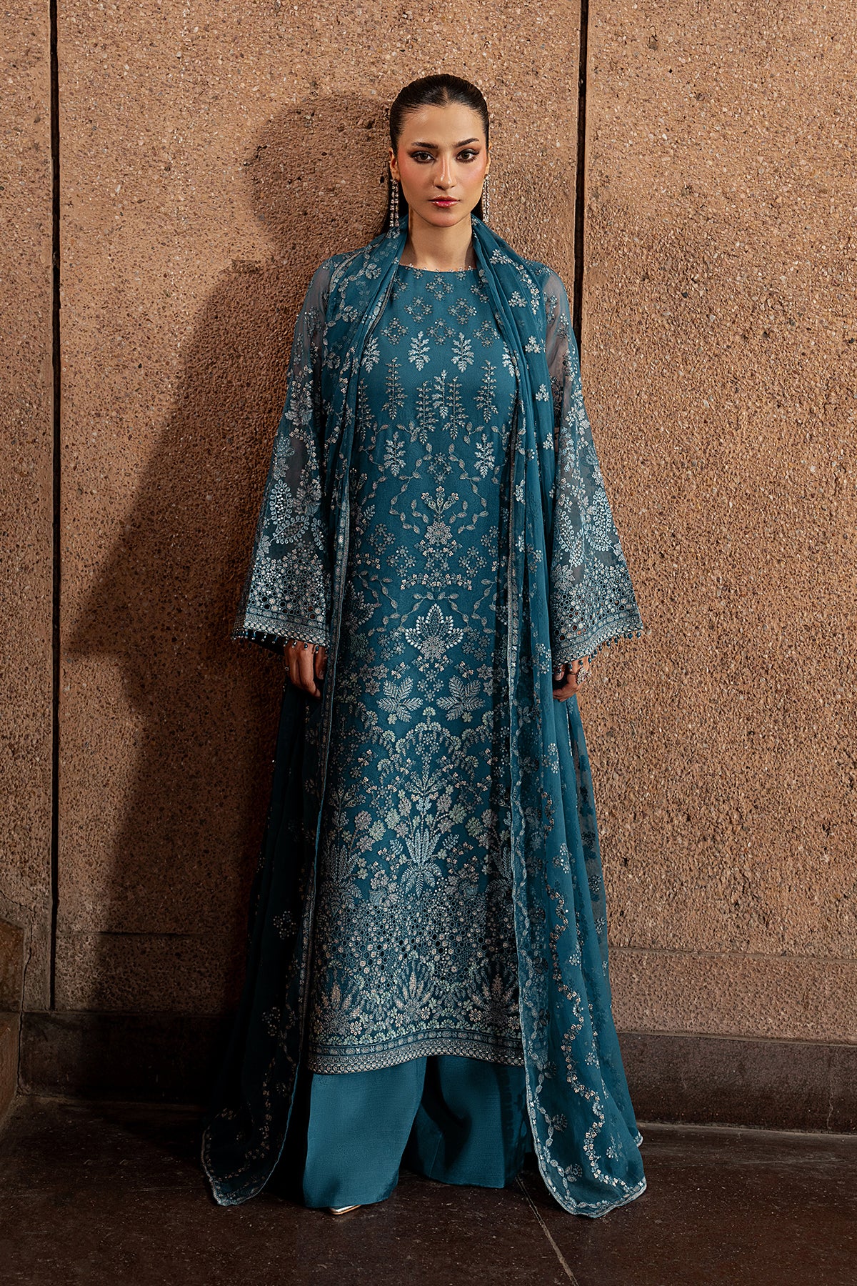 Woman wearing a blue embroidered traditional outfit against a textured wall.