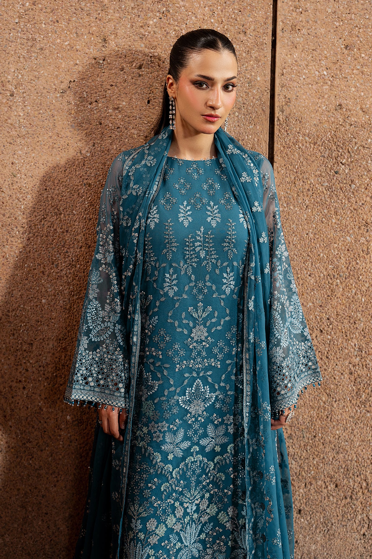 Woman wearing a teal lace dress with a matching shawl against a brown wall.