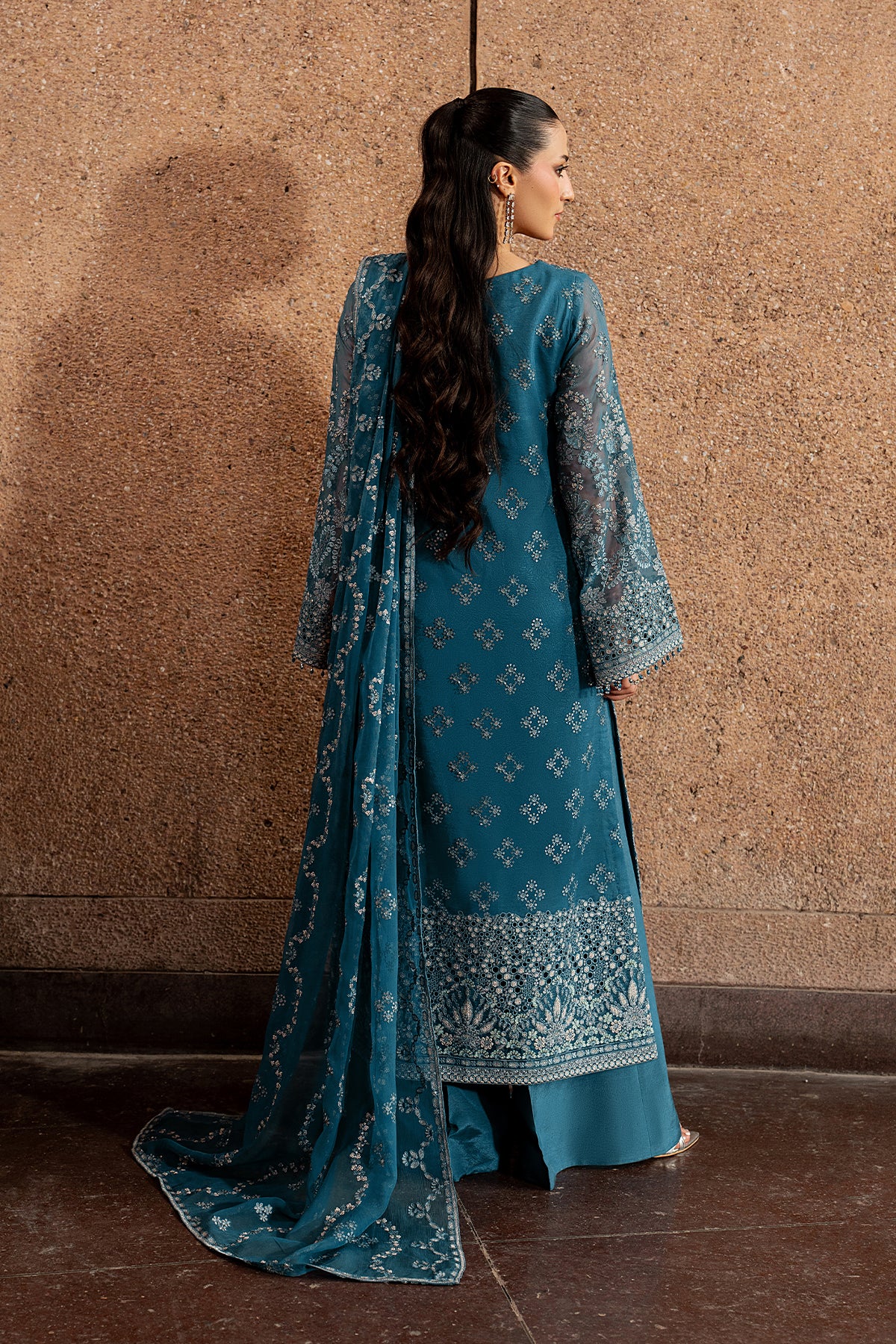 Woman in a blue embroidered traditional outfit standing against a textured wall.