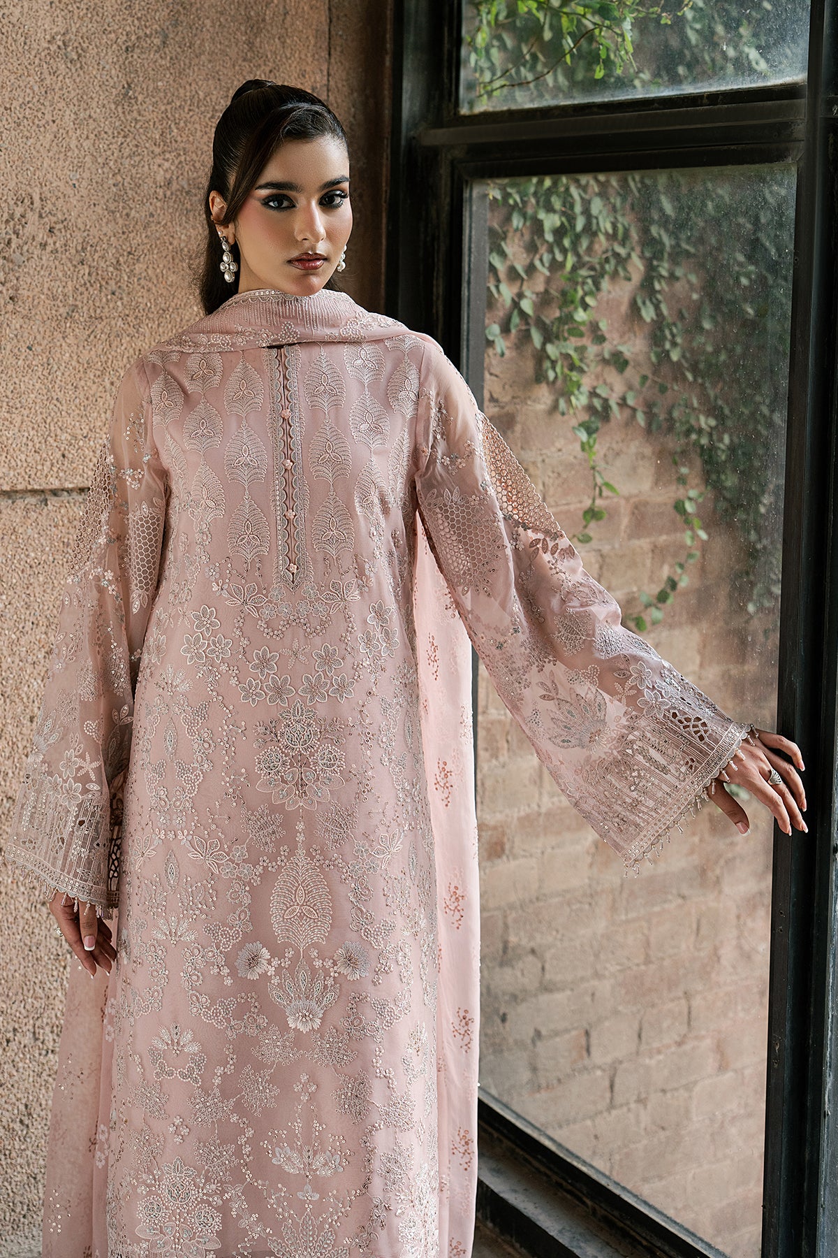 Woman wearing a light pink embroidered outfit standing by a window with greenery outside.